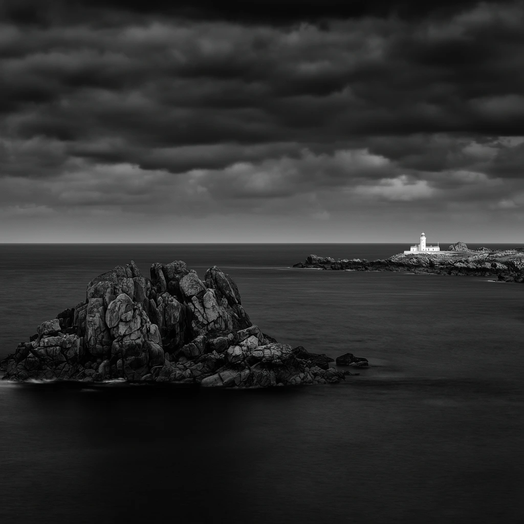 A dramatic black and white panoramic coastal landscape under a heavy, brooding sky. A massive, jagged rock formation deeply textured with crevices dominates the left foreground, rising from a vast expanse of smooth, dark water. In the distant right background, a small, luminous white lighthouse with an adjacent building sits isolated on a rocky peninsula. A crisp, straight horizon line divides the velvety sea from the densely clouded atmosphere above. The composition employs a wide aspect ratio and long exposure photography, rendering the ocean surface as a silky plane that contrasts sharply with the rugged, dark stones and the tiny, brilliant structure.
