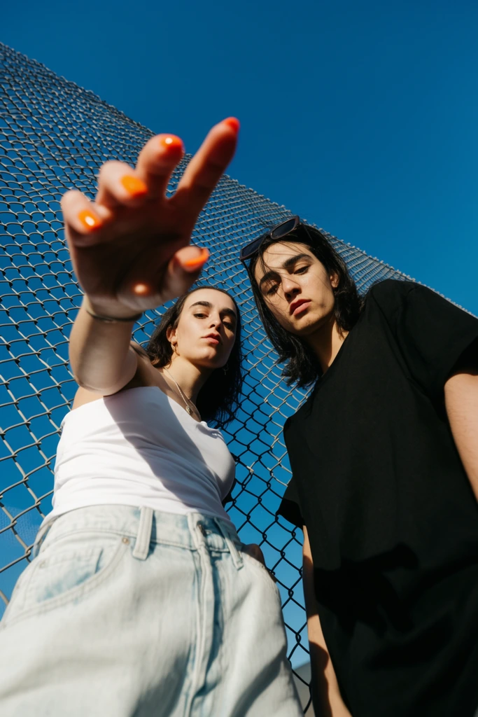 An extreme low-angle view looking up at two figures standing behind a diagonal chain-link fence against a vivid blue sky. A figure on the left in a white top and light denim reaches a dramatically foreshortened hand with bright orange nails toward the lens. Beside them, a person in a black t-shirt with sunglasses resting on dark hair gazes downward. Harsh direct sunlight creates sharp, high-contrast shadows across their bodies, defining a dynamic street photography aesthetic.
