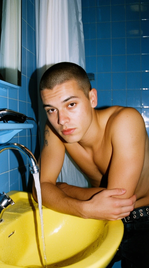 A shirtless young man with a buzz cut rests his forearms on a bright yellow sink, gazing directly at the lens with a neutral expression. Water runs steadily from a curved chrome faucet. He wears dark trousers secured by a grommet belt and has a faint tattoo on his right arm. A pair of black hair clippers sits on a light blue ledge to the left. The background features glossy blue square wall tiles and a partially visible white shower curtain. Harsh direct flash lighting casts deep, sharp shadows, creating a gritty analog film aesthetic with high-contrast color blocking between his warm skin and the cool room.