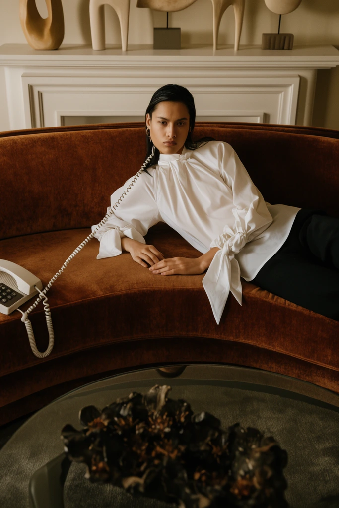 person reclining on a curved rust-brown velvet sofa, crisp white blouse with tied sleeves, white coiled telephone cord, background white mantel with minimalist sculptures, foreground glass table with dark organic decor, warm earthy color palette, moody editorial photography, cinematic lighting