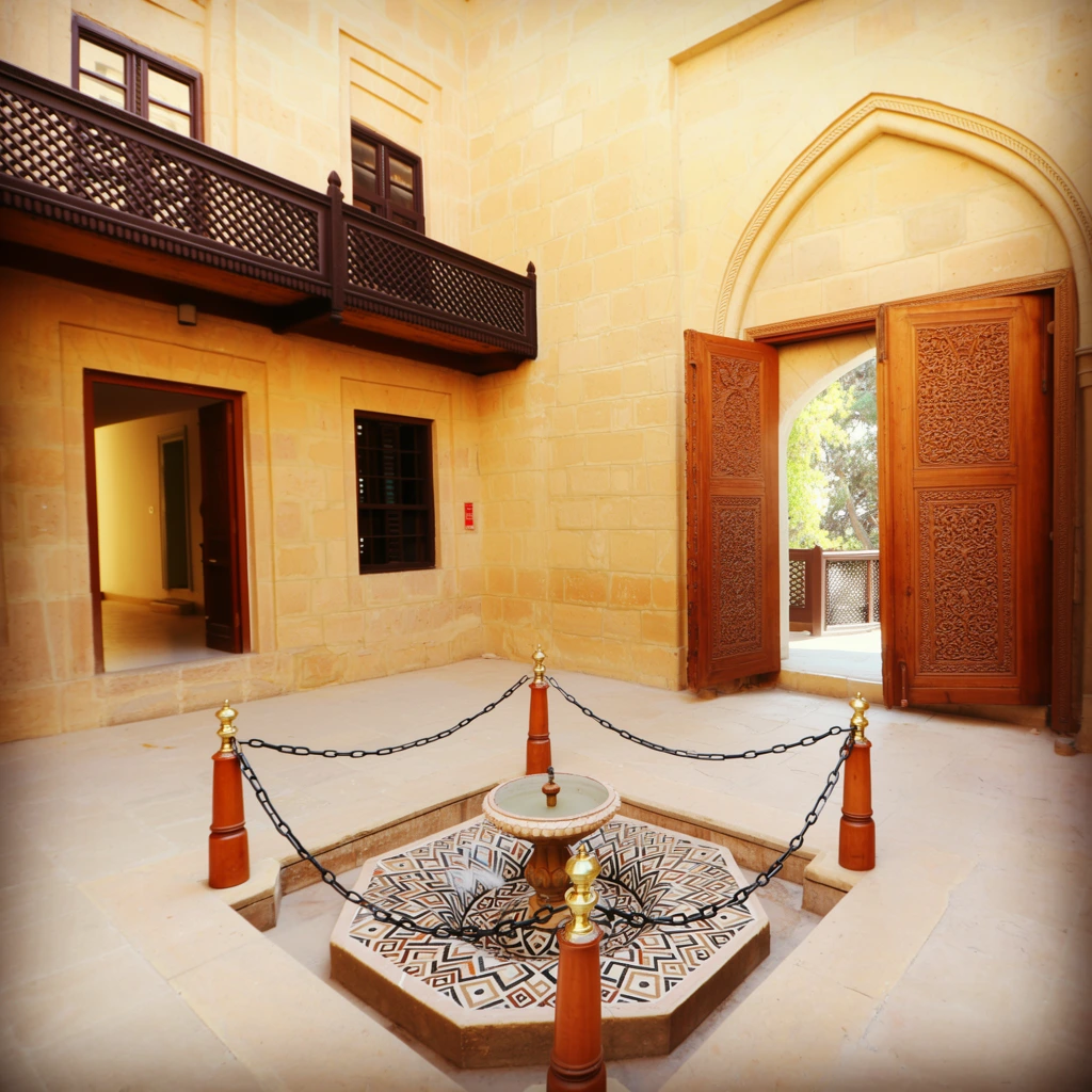 A sunlit sandstone courtyard featuring an ornate sunken fountain with a geometric mosaic basin, bordered by four brass-topped wooden posts linked by a black chain. The warm, pale yellow masonry walls enclose the space, accented by a dark wood lattice balcony and an open doorway revealing an interior hallway. To the right, massive, deeply carved double wooden doors stand open beneath a pointed arch, offering a glimpse of sunlit trees. The eye-level photograph employs a heavy vignette to frame the natural daylight illuminating the smooth stone floors and intricate architectural details.