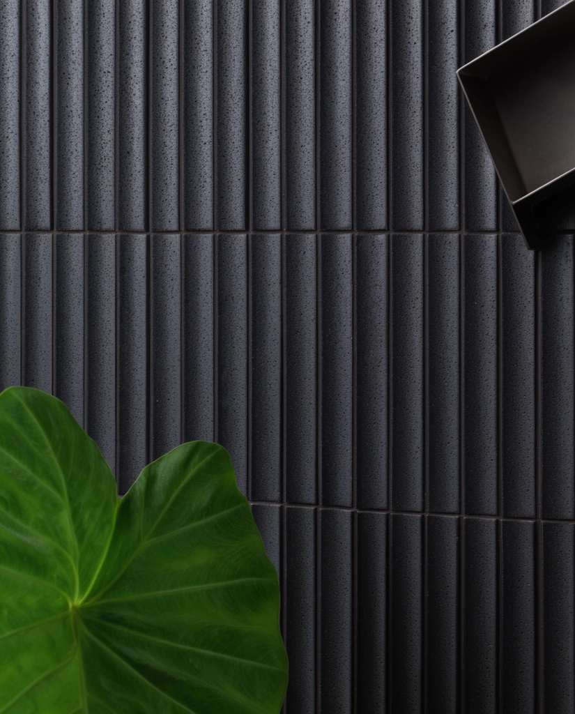 A wall of dark charcoal, vertically fluted ceramic tiles with a porous, matte texture forms a rigid geometric backdrop. Soft directional lighting creates deep vertical shadows within the fluted grooves. In the lower left, a large, vibrant green heart-shaped leaf with delicate veining introduces a striking organic element. A dark, angular metal bracket protrudes from the upper right edge. The minimalist, high-contrast composition emphasizes the tactile interplay between precision industrial surfaces and natural botanical forms.