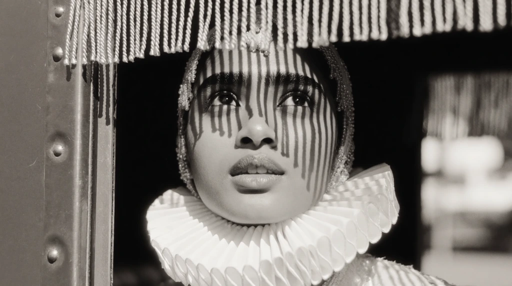 A young South Asian woman with thick dark eyebrows gazes upward with parted lips from inside a riveted metal carriage. She wears an intricate beaded headpiece and a voluminous, stiffly pleated white ruffle collar framing her neck. Thick dangling fringe hangs from the upper window frame, casting striking, high-contrast vertical striped shadows directly across her forehead and face. The tight close-up is framed by the rigid vertical metallic edge of the window on the left. The black and white cinematic photography features a soft vintage film grain and shallow depth of field, blurring the brightly lit outdoor background into abstract shapes.