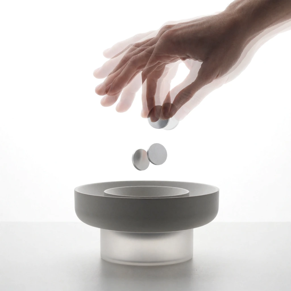 A stroboscopic studio photograph captures the sequential motion of a semi-transparent hand dropping metallic discs into a minimalist gray vessel against a pure white background. Entering from the upper right, the hand is rendered as a multiple exposure with three overlapping, ethereal iterations tracking a downward trajectory. Suspended in mid-air below the fingers, two silver circular tokens fall toward a sleek receptacle. This modern object features a matte gray, shallow outer bowl and a concentric inner depression, resting upon a translucent, frosted white cylindrical base. High-key lighting establishes a pristine atmosphere, casting soft shadows and highlighting smooth gradients on the matte surfaces. The sharply focused, static design object contrasts strikingly with the ghostly, motion-blurred human interaction within the clean, clinical composition.