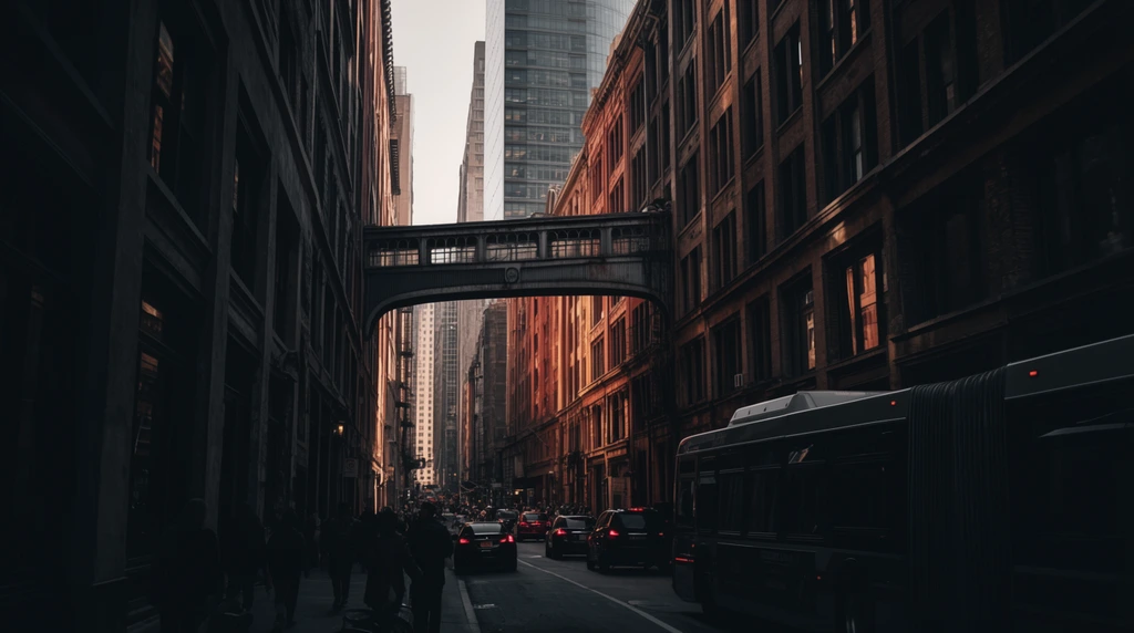 deep urban street canyon, towering historic masonry buildings flanking a narrow avenue, dramatic cinematic lighting, deep shadows contrasting with warm golden sunlight hitting distant building facades, an industrial pedestrian skybridge connecting the structures mid-air, a modern glass skyscraper rising in the background, street level bustling with dark silhouettes of pedestrians, cars with glowing red taillights, a large articulated city bus in the right lane, low-angle one-point perspective drawing the eye down the road, moody color palette of charcoal, rust, and amber, high contrast architectural photography