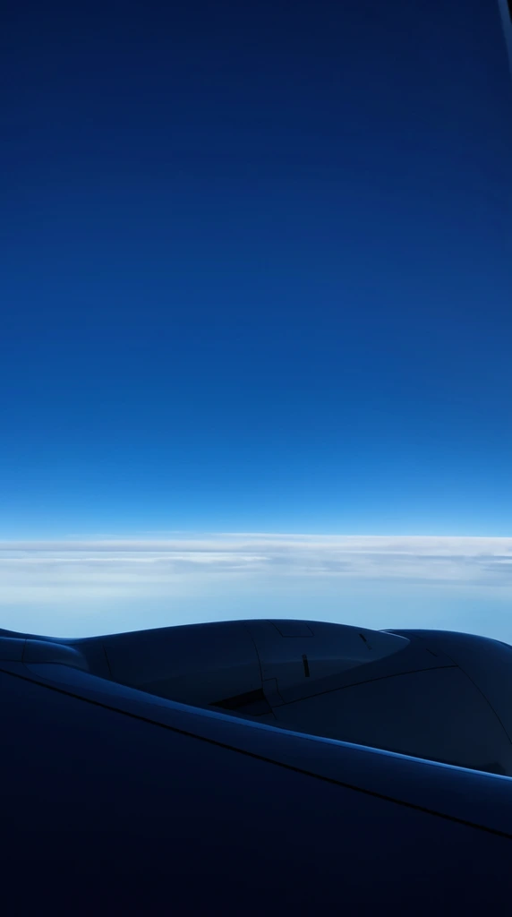 view from airplane window, dark curved wing and engine nacelle silhouetted in foreground, vast deep blue sky filling upper two thirds, smooth gradient transitioning to light azure at horizon, thin horizontal layer of wispy white clouds below, minimalist composition, high altitude aerial perspective, natural daylight, stark contrast between dark metal and bright sky