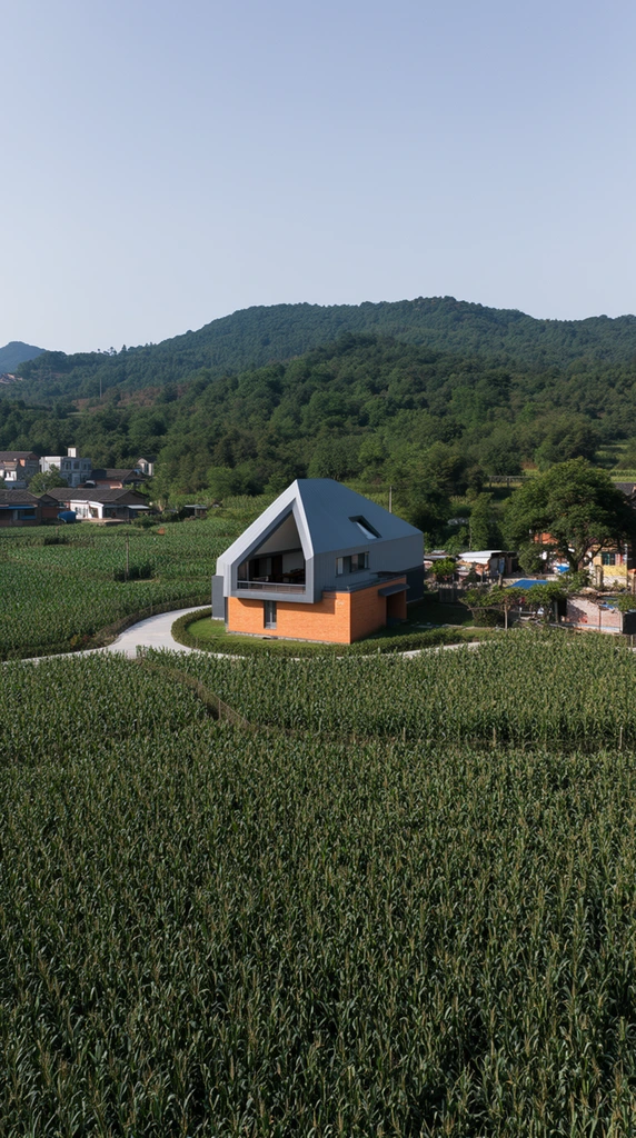 A modern architectural home stands prominently in a vast field of tall green crops. The geometric structure features a striking asymmetrical gray pitched roof sheltering an open second-story terrace, resting on a warm orange-brick lower level. A curved paved pathway winds through the dense agricultural field toward the house. Background rolling hills covered in lush green forests rise against a clear pale blue sky. Scattered traditional rural homes sit to the left, while trees and secondary structures cluster on the right. This elevated wide-angle landscape photography captures the stark juxtaposition of contemporary design against an expansive organic environment under bright natural daylight.