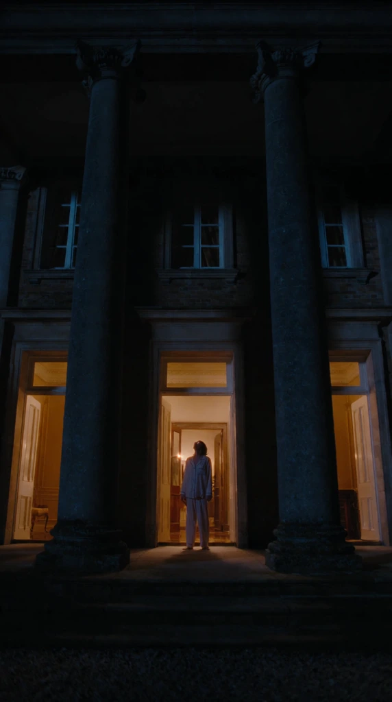 A silhouetted figure in loose pajamas stands in the warmly lit doorway of a dark manor at night. Gazing upward, the person is framed by open double doors and a glowing golden interior. Two massive dark stone columns dominate the foreground, creating a symmetrical architectural frame. Three tall entryways radiate warm light, contrasting sharply with the deep blue exterior shadows. The cinematic low-angle widescreen shot emphasizes the imposing scale of the building and the stark chiaroscuro between the cold outdoor darkness and the inviting indoor glow.