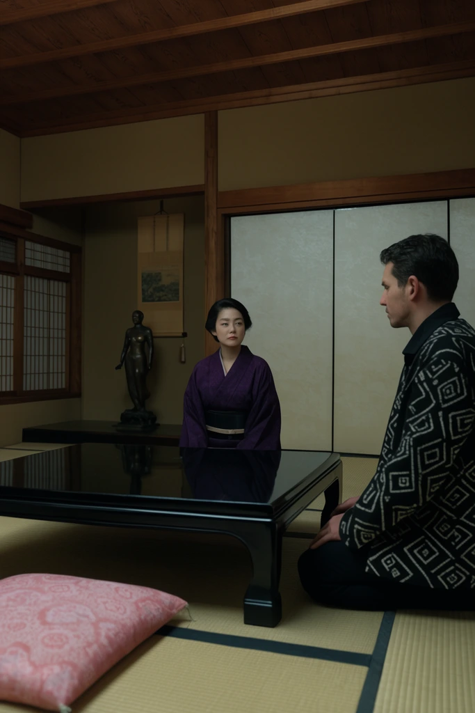 A low-angle cinematic shot of a traditional Japanese interior featuring tatami mats and sliding shoji screens. A woman in a dark purple kimono sits centered behind a polished black low table. To the right, a man in a black jacket with white geometric patterns kneels on the floor, facing her. A pink patterned cushion rests in the left foreground. In the background alcove, a dark bronze figure stands beneath a hanging scroll, illuminated by soft, muted lighting across earth-toned walls.