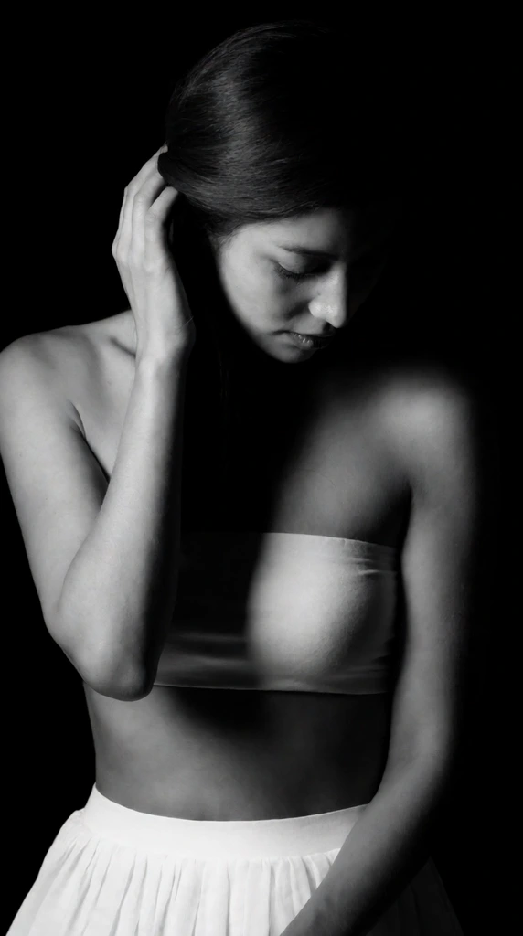 High-contrast black and white portrait of a downward-gazing woman against a pure black void. Wearing a white bandeau and pleated skirt, her hand brushes swept-back hair. Dramatic chiaroscuro lighting sculpts her face and bare shoulders from the left, casting deep shadows in a serene fine art photography style.
