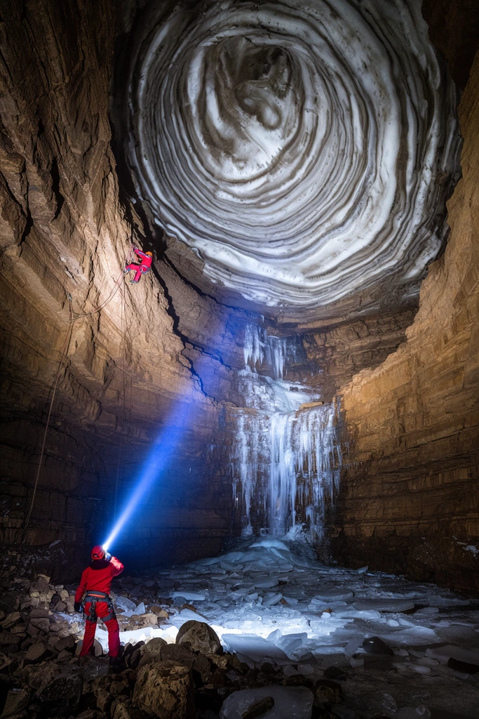 A colossal subterranean cavern featuring layered brown rock walls and a sweeping domed ceiling of swirling white ice strata. In the rocky, ice-strewn foreground, a spelunker in a red suit stands aiming an intense blue headlamp beam diagonally upward, illuminating a frozen waterfall cascading down the central rock face. High on the left wall, a second tiny climber in red is suspended from ropes. Wide-angle adventure photography utilizes dramatic high-contrast lighting to emphasize the massive geological scale of the environment compared to the small human figures.