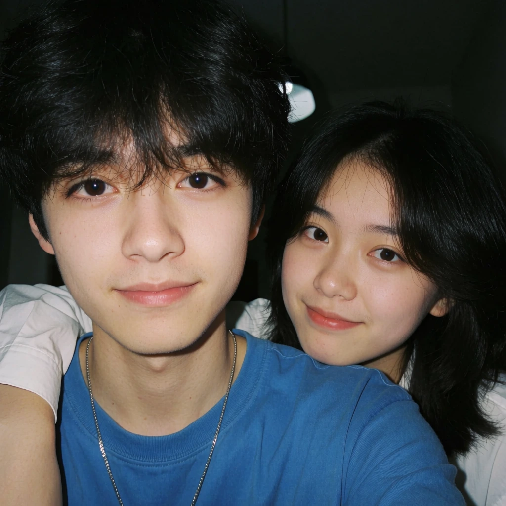 A close, intimate selfie shows two young people indoors at night, their faces filling almost the entire frame, with the person in front smiling softly at the camera while the person behind leans in over his shoulder in a gentle, affectionate pose. digital photography, casual smartphone selfie, candid portrait, contemporary lifestyle portrait, soft flash snapshot, slightly retro social-media aesthetic, naturalistic with mild beautifying softness, cozy, tender, youthful, flirtatious, low-background detail, face-dominant composition, visually simple but texturally rich, adjacent to modern East Asian lifestyle portraiture, faintly reminiscent of film-inspired Instagram photography, warm neutrals, deep blacks, muted skin tones, bright cobalt blue, soft white, peach-pink lips, low-key background, direct frontal flash, moderate contrast, lifted facial highlights, subdued shadows, shallow apparent depth from close focus, likely phone front camera or handheld compact, visible fine grain/noise, slight skin smoothing, subtle flash glare in background, no major motion blur. In the center-left foreground is a young man with pale skin and thick, tousled black hair parted loosely down the middle, several wisps falling over his forehead and near his eyes. He looks directly into the lens with dark brown eyes, relaxed eyebrows, and a small closed-mouth smile. His face is angled slightly leftward, and his head occupies most of the left half of the image. He wears a bright blue crewneck T-shirt covering the lower right half of the frame, and a thin silver chain necklace drops from his neck toward the center bottom. Behind him on the right side is a young woman with similarly dark, softly layered hair framing her face, her head tilted slightly inward toward his left cheek. She also looks at the camera with a calm, affectionate expression and a faint smile. Her face sits just above his right shoulder, close enough that their cheeks nearly touch. On the far left edge, partially obscured, is a white shirt sleeve and part of an arm, likely belonging to the front subject. The background is very dark and indistinct, suggesting an indoor room or nighttime interior; near the top center and top right are a few blurred warm light spots, and a dim reflective object appears along the left edge, but almost all attention is pulled to the two illuminated faces.