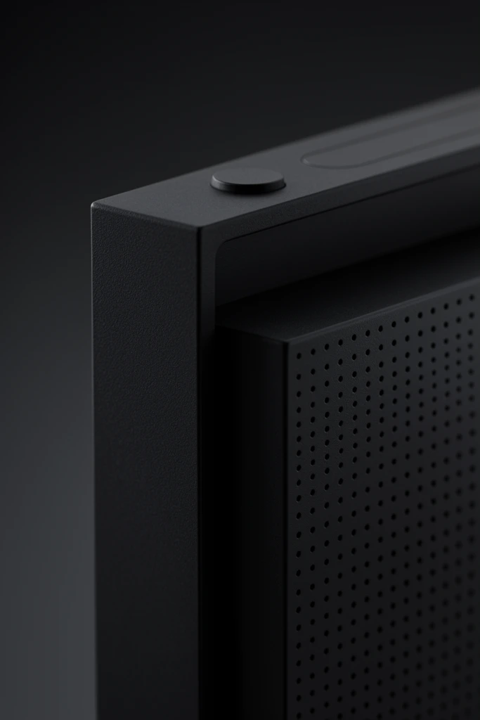 A close-up macro shot highlights the sleek corner of a matte black electronic device against a dark charcoal gradient background. The minimalist geometric structure features a stepped design where a thick outer shell overhangs a recessed inner block. A uniform grid of tiny circular ventilation holes covers the side panel and the recessed surface. Along the prominent upper edge, a small raised circular button sits beside a shallow pill-shaped indentation. Soft low-key lighting casts subtle highlights on the crisp edges, revealing a fine granular texture on the dark, matte material. The shallow depth of field keeps the nearest corner sharply in focus while gently blurring receding lines into the shadowed environment, emphasizing stark industrial design.
