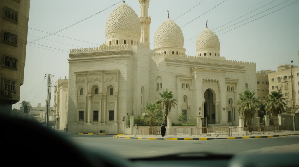 grand light stone mosque with intricately patterned domes and a tall minaret, viewed across an empty urban intersection, blurred dark vehicle dashboard in extreme foreground, hazy daylight sky cut by crisscrossing power lines, paved street with yellow and black painted curbs, palm trees and metal barricades at the entrance, lone pedestrian in dark clothing, flanking multi-story buildings, muted greenish-yellow atmospheric tint, candid street photography