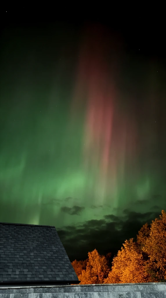 vibrant aurora borealis in a dark night sky, expansive diffuse green glow, vertical reddish-pink light pillars ascending into the darkness, scattered faint stars, dark low-hanging clouds near the horizon, dark gray shingled gabled roof in the lower left, tree canopies vividly illuminated from below by warm golden-orange light in the lower right, gray stone wall at the bottom edge, low-light night photography, high contrast between celestial greens and warm terrestrial oranges