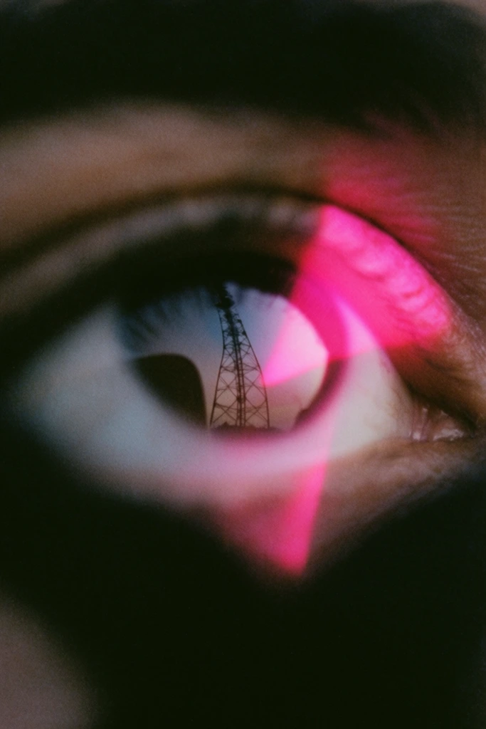 A macro close-up of a human eye reflecting a tall lattice tower, captured with a gritty, lo-fi analog film aesthetic. The central focal point is the glossy curvature of the cornea, displaying a crisp, dark reflection of an intricate steel lattice tower stretching toward a pale, desaturated sky. Heavy film grain, soft overall focus, and deep, crushing black shadows tightly frame the eye and surrounding skin. A striking, intense neon magenta light bleed cuts across the composition, casting a vivid, glowing pink optical flare directly over a section of the iris, sclera, and eyelid. Extreme foreground elements appear as dark, heavily blurred silhouettes obscuring the peripheral edges of the frame, creating dramatic depth and a tightly compressed perspective. The lighting establishes a dim, moody twilight atmosphere, juxtaposing the muted, underexposed skin tones and deep shadows with the harsh, vibrant artificial pink light artifact.