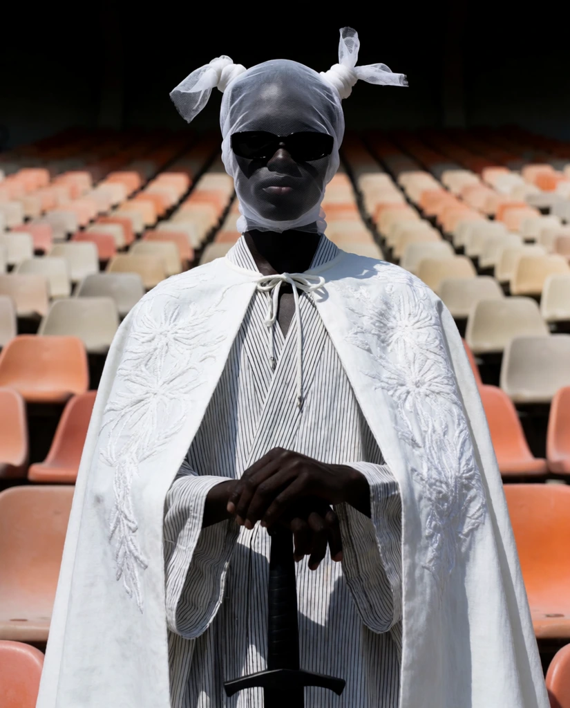 figure in an empty stadium, sheer white mesh headwrap with horn-like knots, dark sunglasses, white embroidered cape over a striped robe, hands resting on a black sword, stark shadows, low angle, rows of faded orange and beige seats, avant-garde fashion photography, symmetrical composition