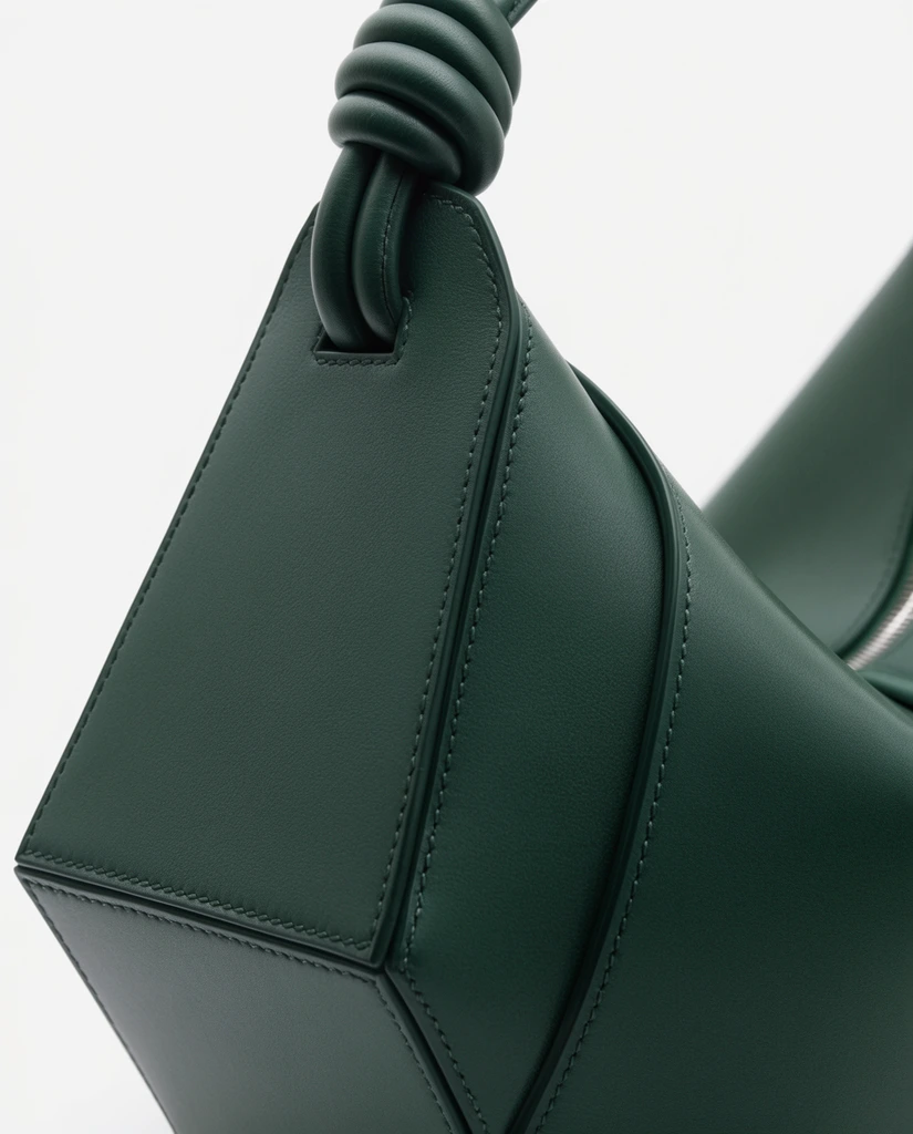 macro product photography of a dark forest green leather handbag, angular structural side panel, smooth matte leather texture, meticulous tonal topstitching tracing geometric edges, tightly knotted leather strap looped at the upper apex, sweeping curved seam on the right, glimpse of a metallic zipper track, isolated on a stark white background, soft diffused studio lighting highlighting volumetric curves, minimalist high-fashion composition