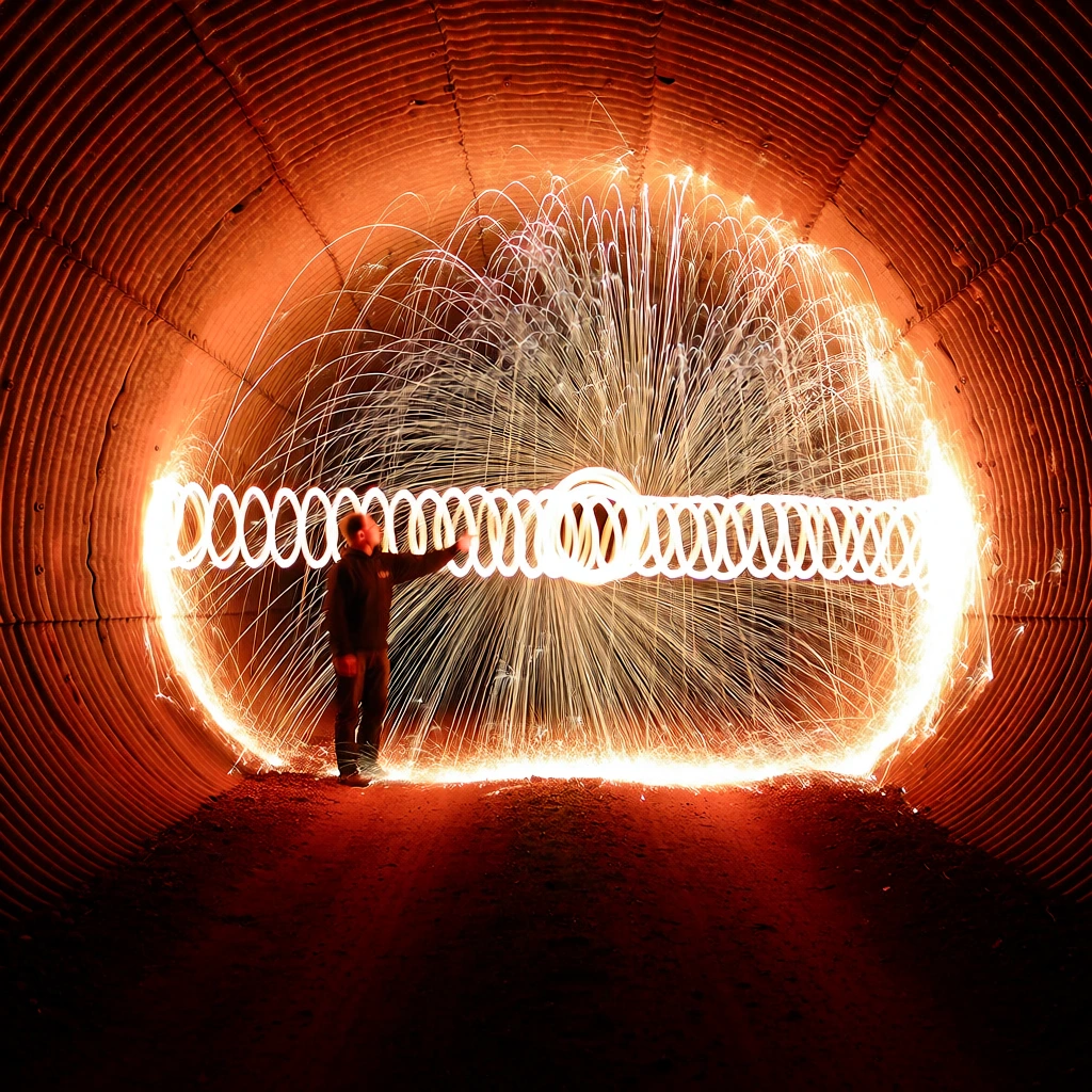 A person stands silhouetted inside a large, circular corrugated metal tunnel, surrounded by a dramatic light painting display featuring a glowing white spiral and a massive burst of radiating sparks. Positioned to the left of the center, the figure wears dark clothing and reaches their right arm toward a horizontal, slinky-like coil of brilliant white light that extends through the middle of the frame. From the center of this coil, a spherical explosion of thin, luminous white and orange streaks branches out in every direction, filling the diameter of the tunnel with intricate, thread-like lines of light. The environment consists of the interior of a ribbed metal culvert, where the horizontal ridges of the walls are illuminated in vibrant shades of orange and deep red by the central light source. The ground is a dark, textured surface of dirt or gravel, heavily shadowed and cast in a crimson glow. The photograph utilizes a long-exposure technique to capture the movement of light, creating a high-contrast composition where the fiery orange and bright white elements pop against the dark, shadowy recesses of the tunnel. The perspective is a straight-on shot from within the tunnel, emphasizing the symmetry of the circular structure and the expansive reach of the light painting.