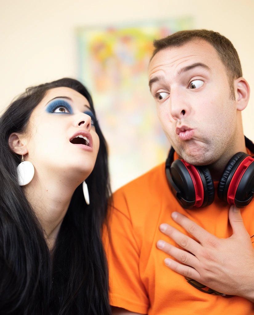Two individuals display exaggerated facial expressions in a warmly lit close-up shot. On the left, a woman with long dark hair, heavy blue eyeshadow, and large white circular earrings tilts her head back, mouth visibly open. Beside her, a man in a bright orange t-shirt stares sideways with wide eyes and pursed lips, pressing a hand against his chest. Bulky black and red headphones rest around his neck. Soft, high-key lighting illuminates their faces against a shallow depth of field. A blurred pale wall with a vibrant, indistinct rectangular artwork forms the background, keeping focus firmly on their contrasting postures.