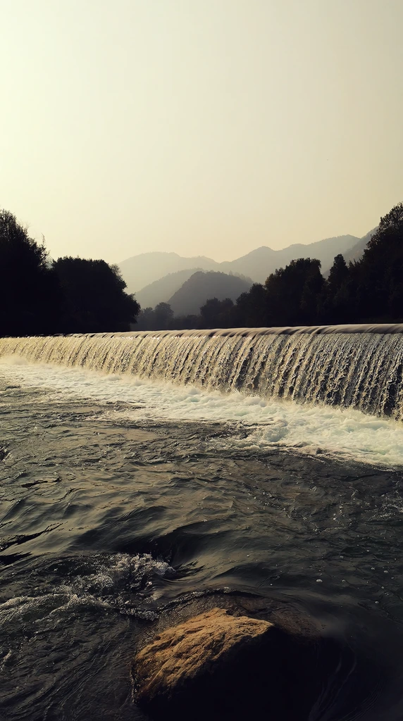 A wide river cascades over a low concrete weir, forming a continuous curtain of rushing white water. Dark rippling currents and a large textured boulder fill the foreground. Silhouetted trees line the riverbanks, fading into hazy rolling mountains in the background. The serene landscape photograph is bathed in soft pale yellow light under a misty sky, emphasizing the contrast between the luminous cascade and the deep shadows of the water's surface.