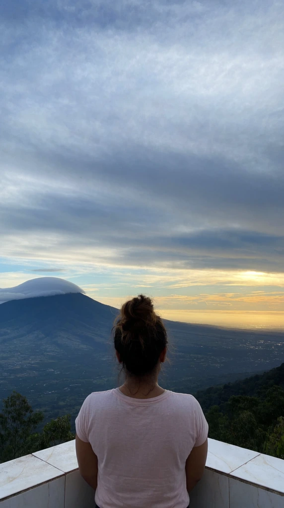 woman with back to camera looking over a vast valley, high messy hair bun, light pink t-shirt, standing at a low white-tiled balcony corner, massive sky filling the frame with streaky blue and gray clouds, golden sunlight glowing on the distant horizon, dark sweeping landscape below, prominent mountain peak on the left capped by a smooth lenticular cloud, wide-angle landscape photography, atmospheric natural lighting