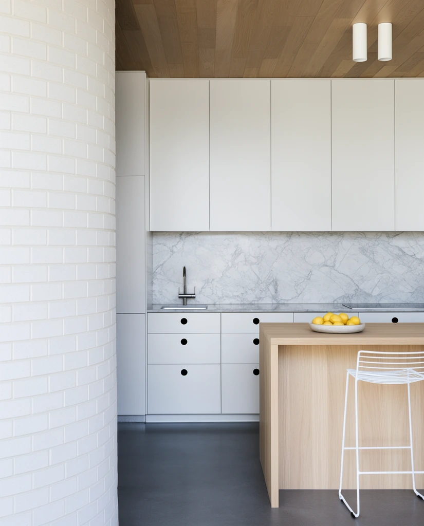 A minimalist kitchen partially obscured by a curved white brick wall in the left foreground. The bright space features matte white cabinetry with black circular cutout handles, a veined marble backsplash, and a wood-paneled ceiling with twin cylindrical lights. A light oak island sits on a smooth dark grey floor, paired with a white wireframe bar stool and a bowl of yellow lemons. Soft diffused daylight emphasizes clean architectural lines and a restrained color palette.