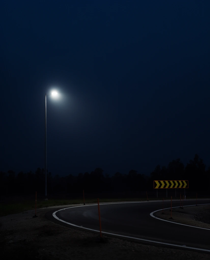 A solitary street lamp illuminates a curving paved road beneath a vast, inky blue night sky. The tall pole on the left shoulder emits a bright, hazy white glow, creating stark contrast against the darkness. The asphalt road sweeps right, bordered by crisp white lines and thin orange delineator poles. A rectangular reflective sign on the outer curve displays repeating yellow and black geometric chevrons. A silhouetted treeline forms a low horizon swallowed by heavy shadows. The photographic composition utilizes extensive negative space in the upper two-thirds, enhancing the moody, liminal atmosphere through low-key lighting and deep shadow tones.