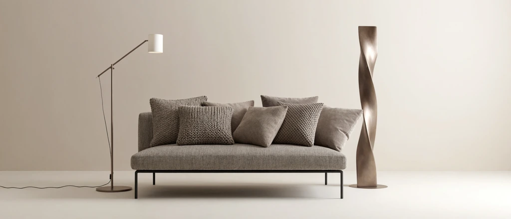A minimalist interior vignette featuring a low-profile heathered grey sofa resting on a thin black metal frame against a monochromatic beige backdrop. The seat is layered with square throw pillows in muted taupe and ash tones, showcasing contrasting tactile surfaces like chunky knits, dotted fabrics, and smooth velvet. Beside the sofa stands a tall, twisted brushed-bronze floor sculpture and a slender metallic floor lamp with an angled arm and a white cylindrical shade. A thin black power cord trails across the smooth off-white floor, which transitions seamlessly into a plain, softly illuminated wall under warm, diffused studio lighting.