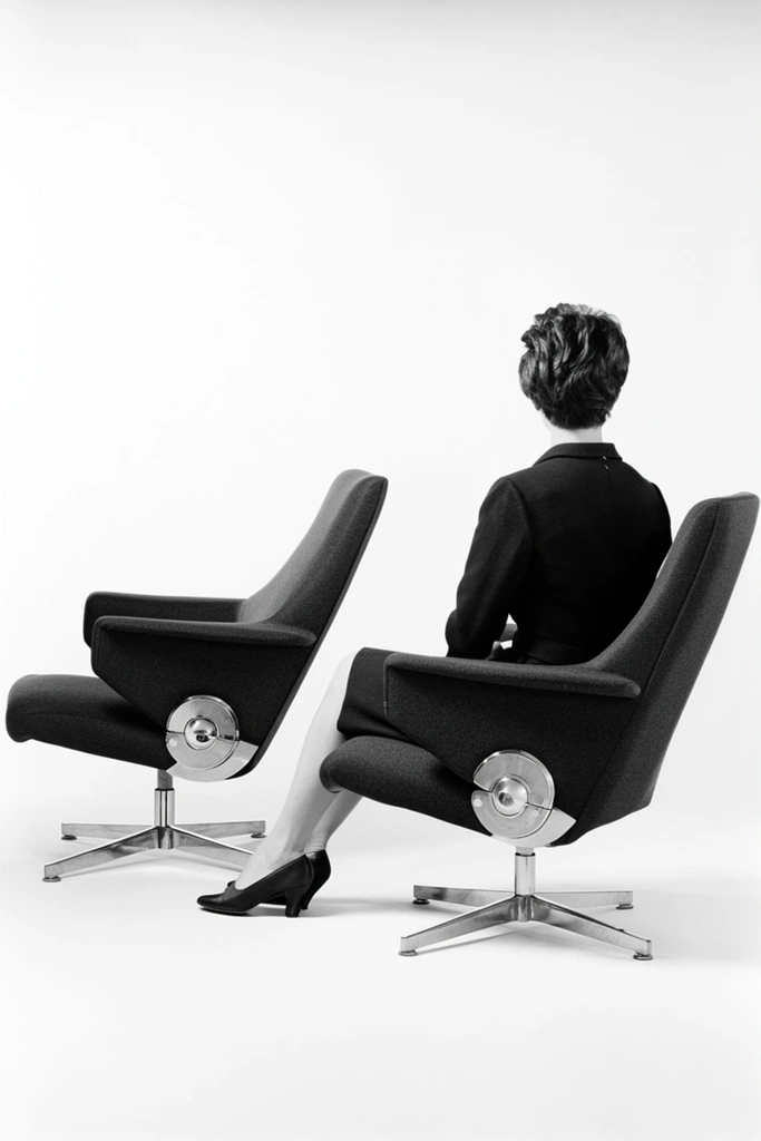 black and white studio photography, two identical dark upholstered swivel chairs with high angled backs and integrated armrests, prominent circular metal pivot hinge on the side, polished four-star metal cross base, a person seated in the right chair facing away from the camera towards the empty left chair, person wearing a dark tailored dress and dark heels with short textured dark hair, stark featureless white background, high contrast lighting, minimalist mid-century modern product staging