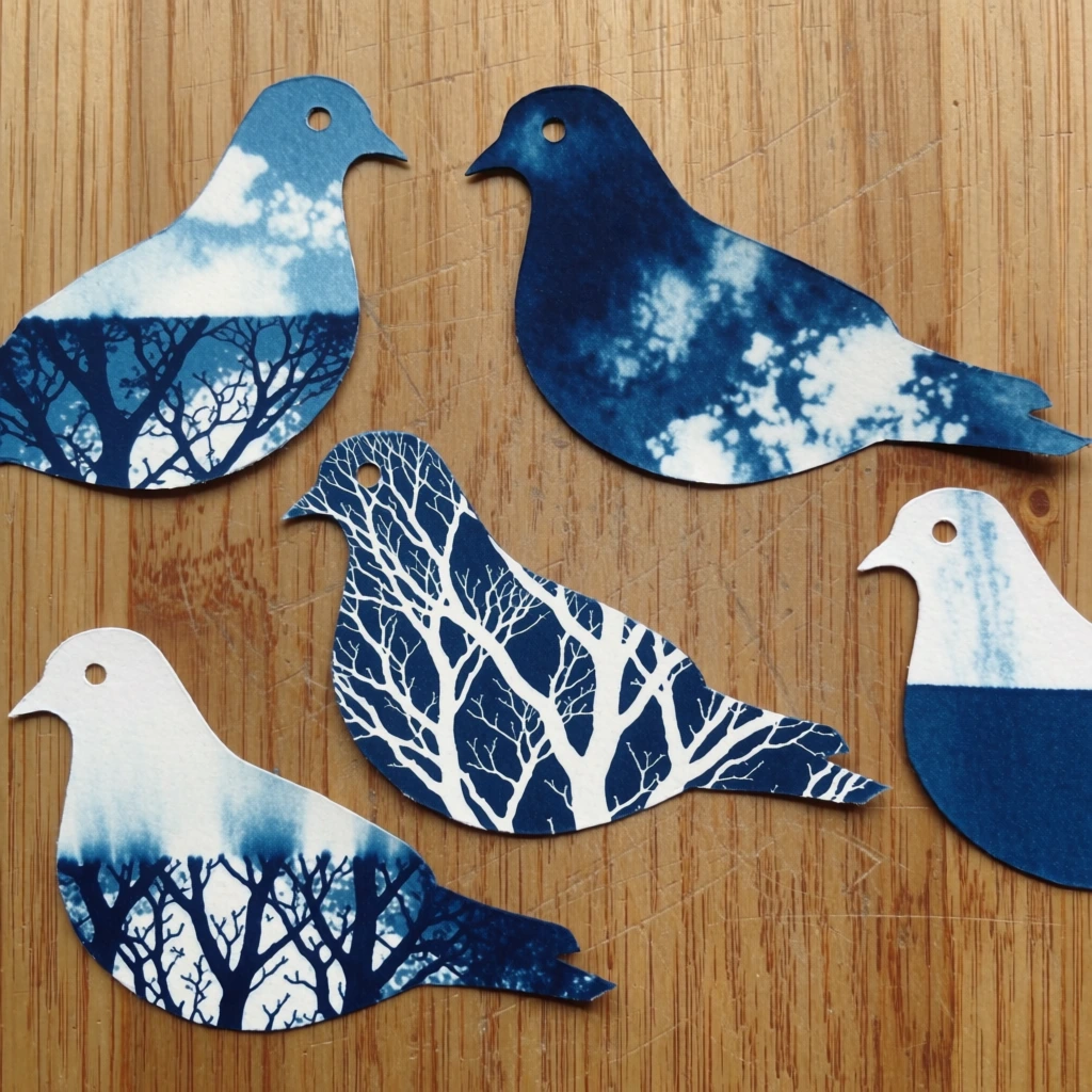 Five bird-shaped paper cutouts featuring cyanotype prints of tree branches are scattered across a light-colored wooden surface in a top-down flat lay composition. The cutouts, resembling the silhouettes of pigeons or doves, vary in their internal patterns; some are deep Prussian blue with white cloud-like textures, while others are off-white with intricate, dark blue silhouettes of leafless tree branches. Each bird cutout has a small circular hole punched in the head to represent an eye. In the top left, a bird profiles to the right with a pale blue upper body and a dark, branch-filled lower half. To its right, a larger bird faces left, rendered in a saturated dark blue with mottled white textures. Centered below these is a bird facing right, showcasing a dense network of fine, blue winter branches against a white background. At the bottom left, a similarly patterned bird faces left with its tail pointing towards the bottom edge. On the far right, a fifth bird faces left, featuring a white upper half that transitions into a dark blue tree line at its base. The background consists of a vertical-grain wooden board, possibly bamboo or plywood, with warm tan and light brown tones, marked by occasional dark scuffs and natural wood grain patterns. The lighting is even and diffused, casting faint, soft shadows beneath the edges of the paper cutouts, which emphasizes their flat, two-dimensional nature against the textured wood. The overall color palette is limited to shades of cyan blue, white, and the natural ochre of the wood.