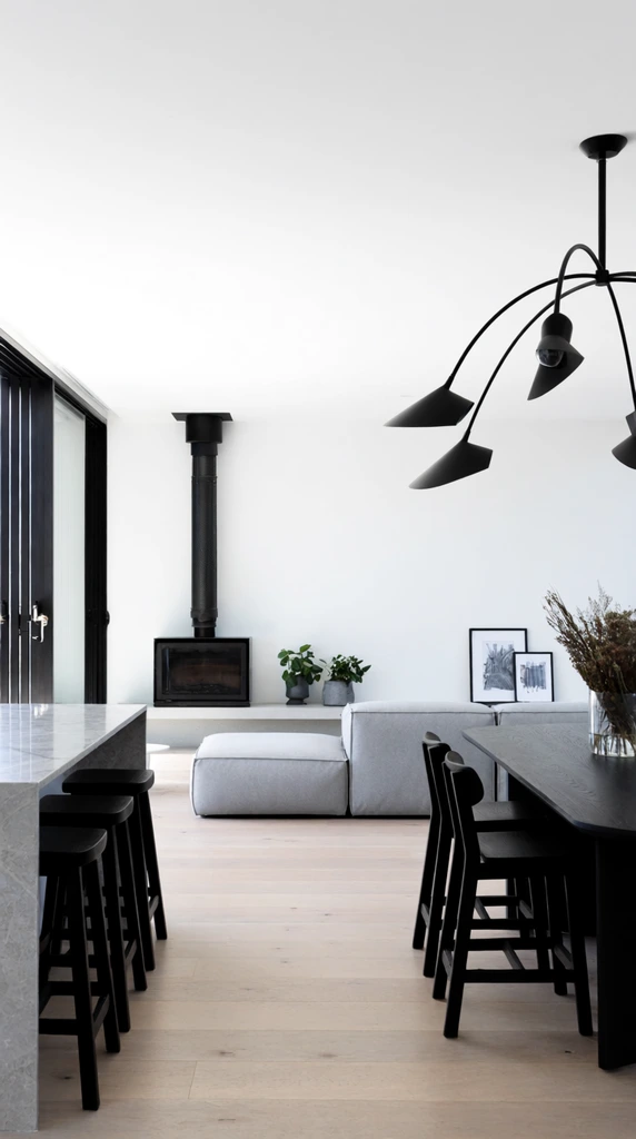 A bright, minimalist open-plan living space illuminated by large black-framed sliding glass doors on the left. A light grey marbled kitchen island with three black wooden bar stools sits beneath a striking black chandelier with sweeping arched arms. Nearby, a black dining table holds dried botanicals in a glass vase. Light wood floors flow toward a low-profile grey modular sofa facing a crisp white wall. A black freestanding wood-burning fireplace with an exposed vertical flue rests on a low continuous hearth alongside small potted plants and framed artwork, establishing a stark, high-contrast contemporary interior.