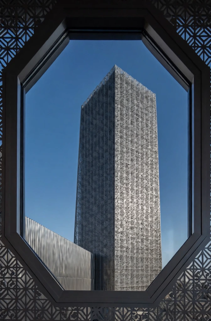 a modern architectural vista viewed through a dark ornate octagonal lattice window, a tall sleek skyscraper ascending into a clear cerulean sky, the building wrapped in an intricate metallic mesh skin with geometric perforations, high-contrast sunlight catching the steel textures, minimalist frame-within-a-frame composition, sharp focus on architectural details and mesh patterns, cool slate and brilliant blue color palette, sophisticated monolithic aesthetic, clean lines