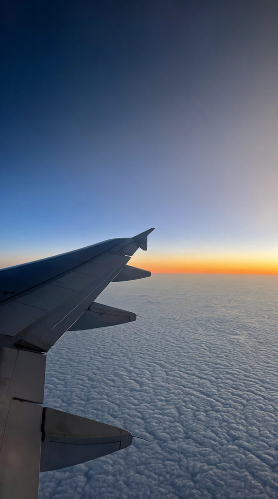 sleek metallic airplane wing with winglet extending from bottom left, vast sea of textured rippled clouds below, smooth sky gradient transitioning from deep navy blue to vibrant sunset orange at the horizon, high-altitude aerial photography
