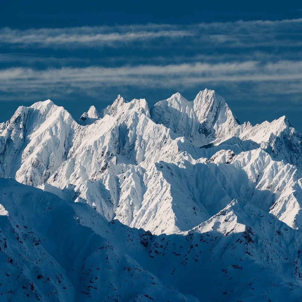 A majestic snow-capped mountain range with jagged peaks and steep ridges dominates the landscape. Brilliant white snow illuminates sunlit upper elevations, contrasting sharply with deep, cool blue shadows pooling across the lower slopes. Captured with a telephoto lens, the intricate layers of the rugged alpine topography are compressed into a dense composition. Above the sharp alpine silhouettes, a deep azure sky is striated with delicate horizontal bands of wispy white clouds, enhancing the crisp, high-contrast, cool-toned aesthetic of the dramatic landscape photography.