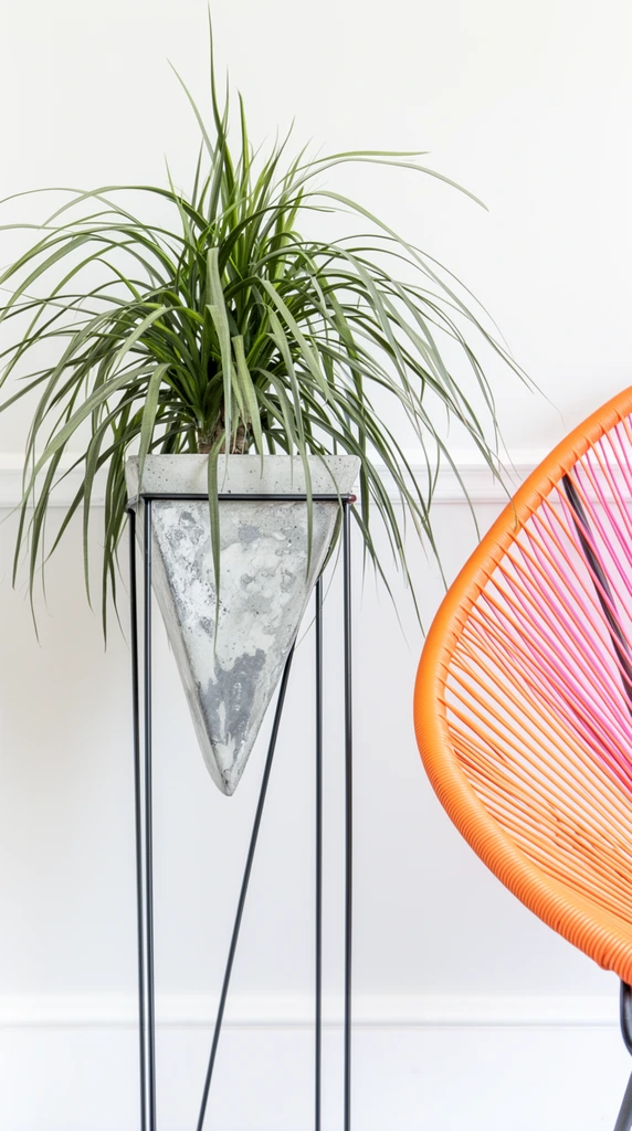 spiky green plant with long cascading leaves, inverted triangular concrete planter featuring a mottled gray texture, tall black metal wire frame plant stand, vibrant orange and pink woven cord lounge chair partially visible on the right, smooth white wall background with a single horizontal molding line, bright diffused natural lighting, minimalist modern interior photography, high-key color palette, clean geometric composition