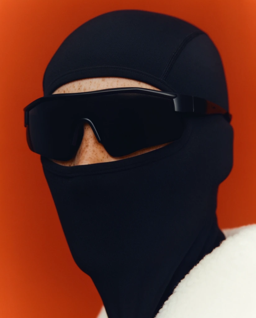 A close-up portrait of a person wearing a tight black balaclava and large opaque wraparound sports sunglasses against a solid red-orange background. The matte fabric covers the head and neck, exposing only a narrow strip of freckled skin across the nose. A thick white textured collar appears at the lower right. The image uses a minimalist fashion editorial photography style with sharp focus and flat studio lighting, creating bold color blocking between the deep black accessories and the vibrant warm backdrop.