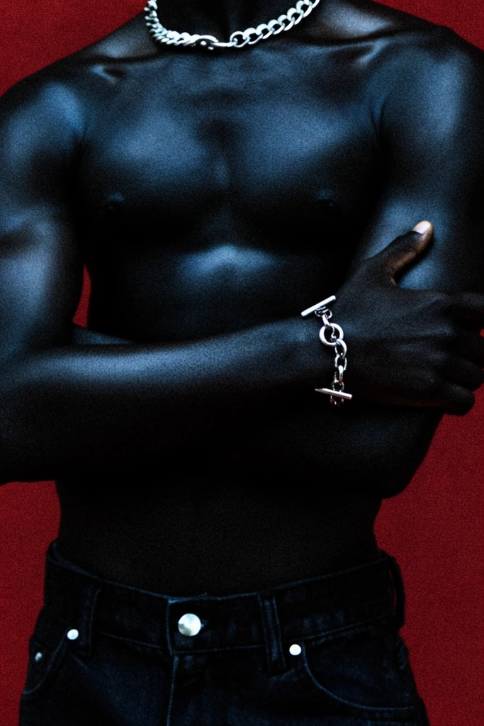 A close-up editorial portrait of a shirtless person with deeply pigmented dark skin, posing with crossed arms against a heavy-grained, deep crimson background. The framing focuses on the muscular torso, cropped just above a thick silver chainlink necklace. A matching silver toggle bracelet adorns the right wrist resting on the bicep, while dark denim jeans are fastened by a reflective silver button. Intense, high-contrast lighting casts cool, bluish highlights across the skin, emphasizing sculptural anatomy. The composition features a raw, cinematic aesthetic defined by dense film grain and a striking color palette of absolute blacks and rich reds.