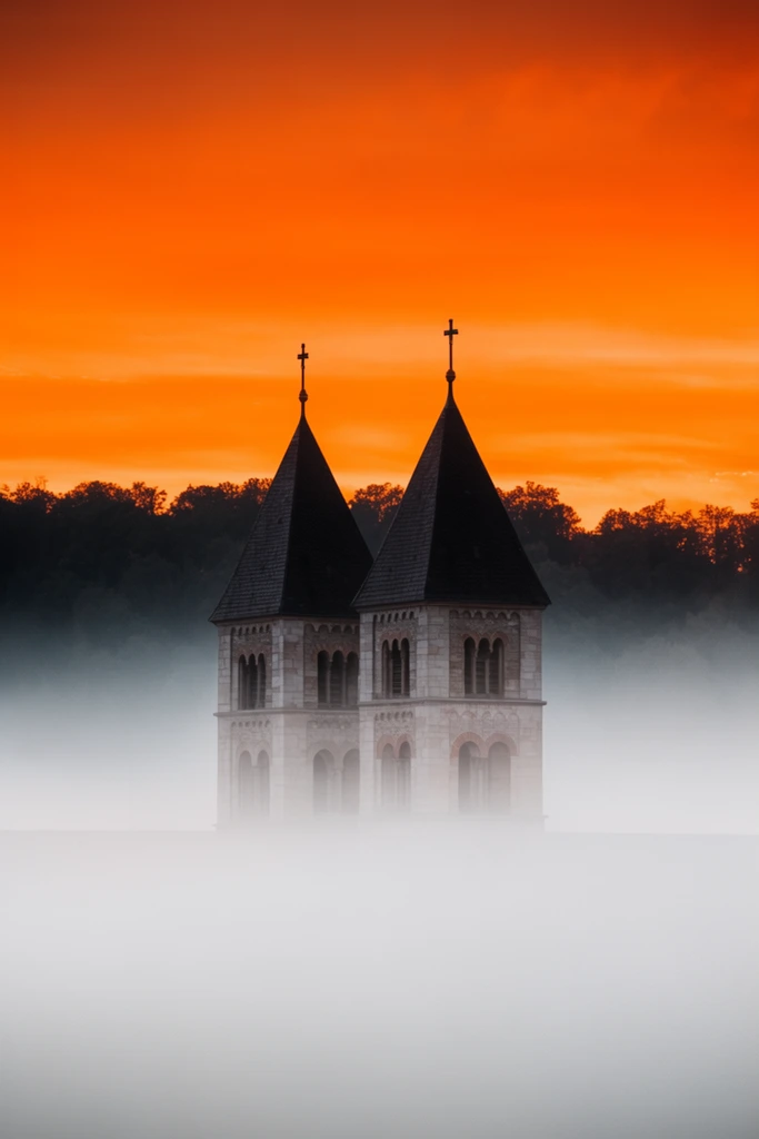 Twin stone church towers with dark pyramidal roofs emerge from a dense sea of smooth white fog. A dark silhouetted treeline stretches behind the structure against a vividly saturated, luminous orange sunset sky.