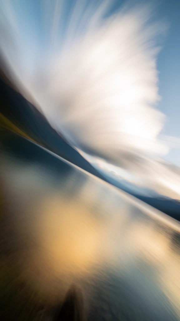 high-key ethereal landscape of a lake reflecting mountain ridges and a massive cloud, intentional camera movement creating a dynamic radial zoom-burst motion blur, soft-focus impressionistic aesthetic, color palette of hazy cerulean blue and luminous milky whites with golden light accents, dreamy and fluid composition where land and sky bleed together in streaks of movement, airy lighting, painterly texture, minimalist yet energetic vibe, cinematic and abstract photography.