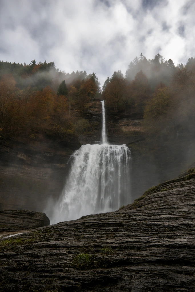A colossal two-tiered waterfall cascades down a sheer, dark cliff. The narrow upper stream impacts a rocky ledge, fanning into a broad, turbulent lower cascade that generates a thick cloud of white mist. Heavily textured dark grey bedrock with prominent horizontal striations and sparse tufts of green grass dominates the foreground. Crowning the cliff is a dense forest of evergreens and autumn-colored trees in rust and gold. Low-hanging fog weaves through the canopy, merging with the waterfall's spray under an overcast sky of textured grey and white clouds. The low-angle perspective emphasizes the immense scale and atmospheric, moody quality of this natural landscape.