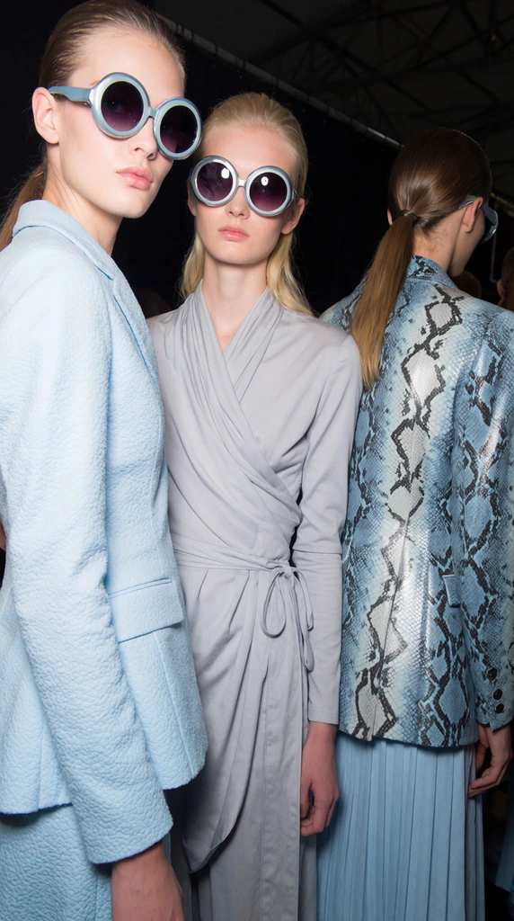 fashion models backstage wearing oversized round futuristic sunglasses, textured pastel blue tailored jackets, blonde model in a draped grey wrap top, model turned away in a blue and silver snakeskin jacket, bright direct flash photography, dark industrial background, cool pastel color palette
