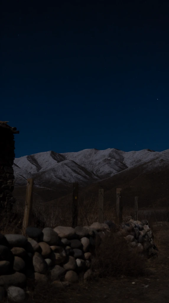 A snow-dusted mountain range stretches across a deep indigo night sky speckled with faint stars. A low rounded-stone wall, wooden fence posts, and dry brush dominate the foreground, leading toward dark foothills. A shadowy stone structure edges the left frame. The low-light, grainy photographic style captures a desolate nocturnal wilderness.