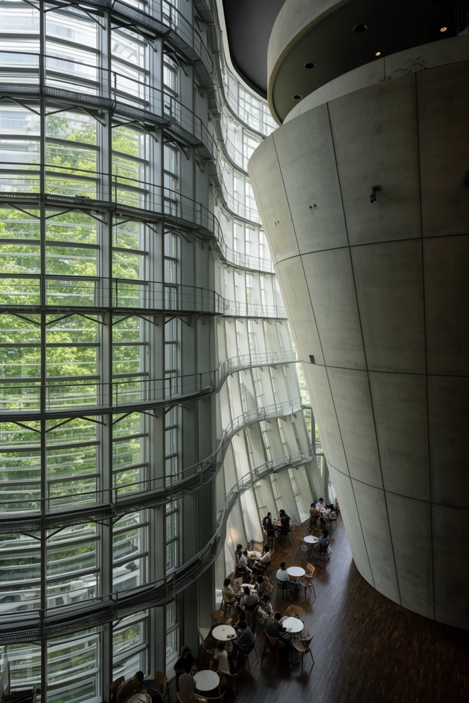 A high-angle view of a sweeping modern atrium defined by an undulating glass curtain wall and massive inverted concrete cones. Lush green trees outside filter daylight into the monumental space. Below, people gather at small round cafe tables scattered across a dark wooden floor, highlighting the dramatic contrast between the colossal brutalist forms and the towering steel-framed glass facade.