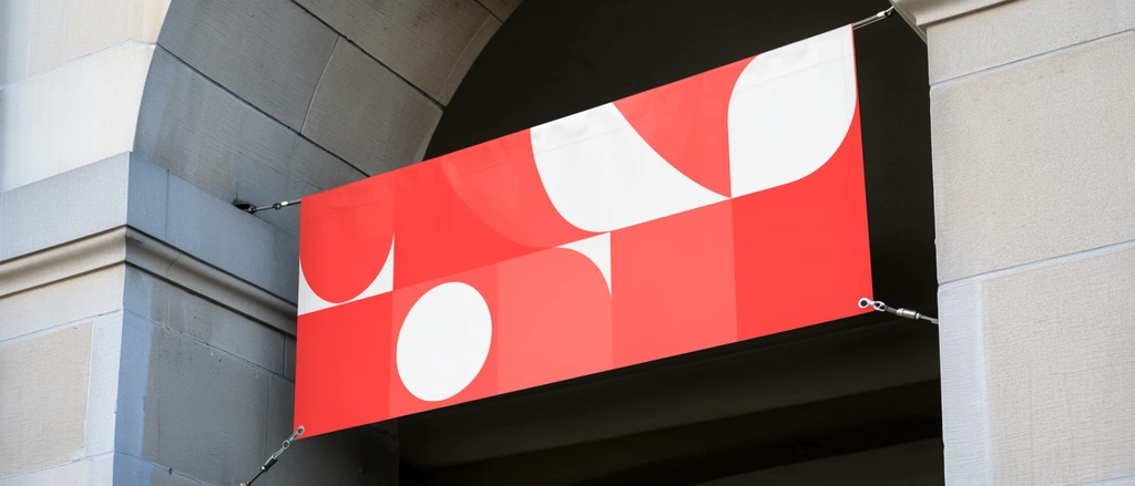 Rectangular banner suspended by metallic tension cables inside a stone archway, abstract geometric graphics in vibrant red coral and white, light gray ashlar masonry, curved classical architecture, low angle perspective looking upward, architectural mockup photography, natural daylight, sharp focus