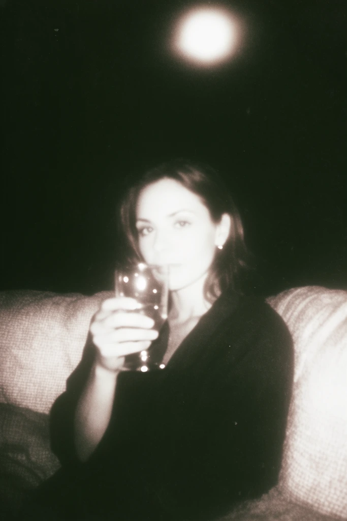 A woman seated on a couch holding a glass, captured in the stark, high-contrast aesthetic of lo-fi vintage flash photography. A harsh, direct, blinding frontal illumination completely blows out the warm, sepia-tinted white highlights on her skin and the heavily textured fabric of the couch, abruptly falling off to plunge the surrounding environment into deep, crushing, inky blackness. The glass in her hand and her gaze catch the intense light, producing sharp, glowing, pinpoint specular reflections that pierce the heavy shadows. An intensely overexposed, hazy circular light orb with a soft glowing halo hangs suspended in the vast pitch-black background overhead. The entire frame is heavily degraded with a dense layer of gritty film grain, murky shadow gradients, soft-focused edges, and a desaturated, monochromatic visual treatment characteristic of expired snapshot film, creating a voyeuristic and deeply atmospheric visual tension.