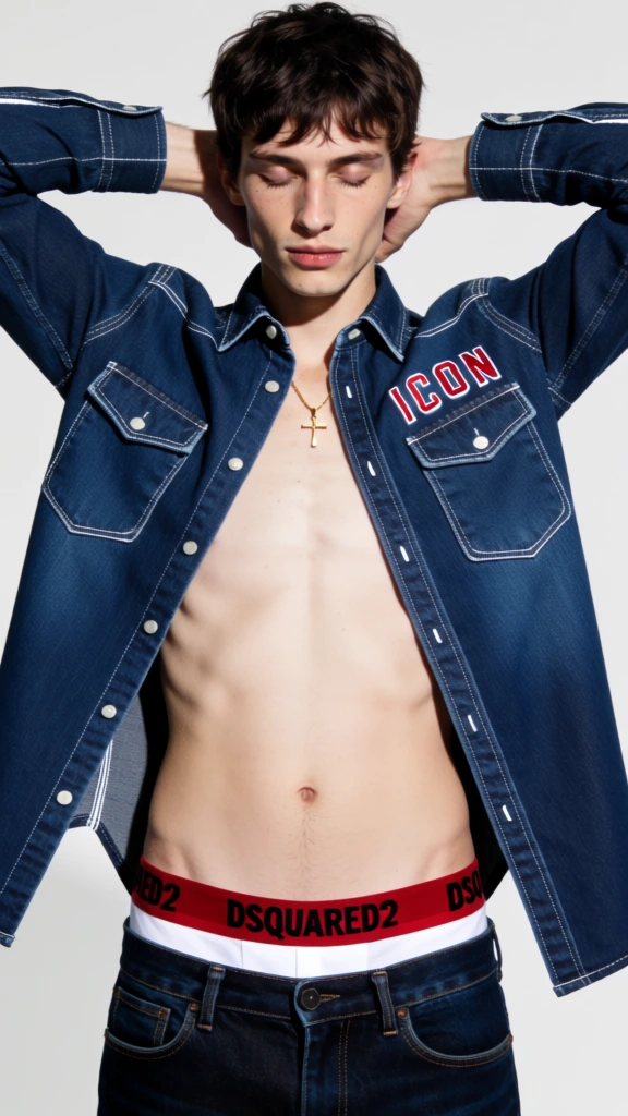 A young man with tousled dark brown hair poses against a light, neutral background, wearing an unbuttoned dark denim shirt and matching jeans. He has a lean physique and pale skin, with his eyes closed and his head tilted slightly downward in a neutral expression. His arms are raised, with his hands resting behind his neck, pulling the dark indigo denim shirt open to expose his bare chest and torso. Around his neck, a thin gold chain holds a small cross pendant. The denim shirt is detailed with contrast white stitching, two chest pockets, and white buttons. On the right chest pocket from the viewer's perspective, the word "ICON" is embroidered in a bold, red, italicized font. His low-slung jeans reveal the waistband of white underwear, which features the brand name "DSQUARED2" in black capital letters and a thin red horizontal stripe along the top edge. The lighting is bright and uniform, creating a high-contrast look between the dark fabric and the pale subject. The composition is a vertical medium shot, focusing on the subject from the waist up against a clean, shadowless backdrop.