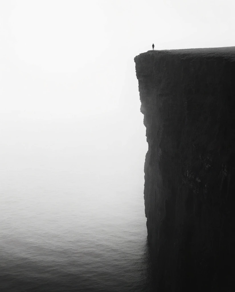 massive dark vertical sea cliff, tiny human silhouette standing on the extreme top edge, stark black and white photography, thick white mist filling the vast negative space on the left, gently rippled ocean at the base, minimalist composition, immense scale, dramatic sheer drop, high contrast