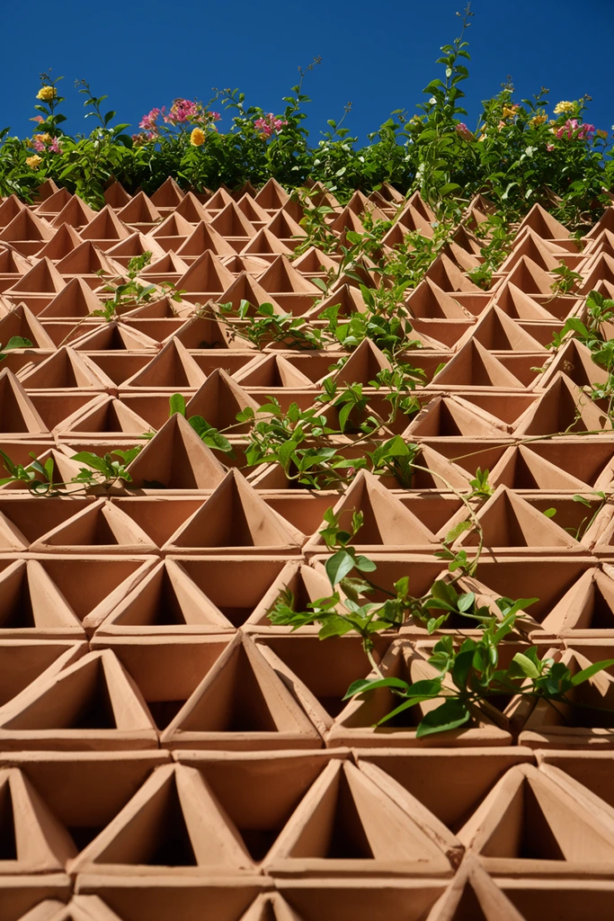 A steeply angled architectural photograph looking upward at a textured facade constructed from repeating matte terracotta modules. The geometric structure forms a grid of hollow diamond-shaped cells crowned by protruding triangular pyramids, creating a sharply jagged surface. Vibrant green climbing vines and leafy stems weave through the geometric voids and cascade over the clay edges, accented by small pink and yellow blossoms near the upper ledge. Bright, direct sunlight illuminates the warm earth tones, casting deep, sharp triangular shadows that emphasize the three-dimensional depth of the screen. A clear, deep blue sky stretches across the uppermost edge, providing a stark cool contrast to the sunlit clay and vivid foliage.