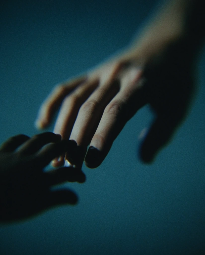 A close-up of a prominent hand reaching downward, interacting with smaller, silhouetted hands appearing near its fingertips against a plain background. The primary hand is rendered with muted warm skin tones, sculpted by stark, directional side lighting that plunges the contours and underside into deep, inky black shadow. The diminutive hands cluster near the extended fingertips as heavily darkened, obscured silhouettes. The plain background is bathed in dense, moody tones of deep teal and dark cyan. The framing utilizes an incredibly shallow depth of field, pinning sharp optical focus solely on the main hand's fingertips while the surrounding elements and edges dissolve into heavy optical blur and thick bokeh. The visual execution is distinctly cinematic and neo-noir, characterized by heavy analog film grain, high-contrast chiaroscuro lighting, and a heavily stylized cold blue and cyan color grade reminiscent of low-light analog photography.